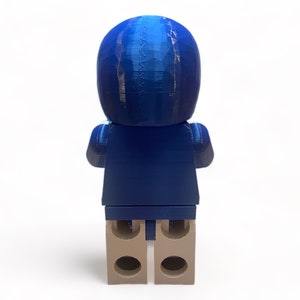 May include: A blue figurine resembling a person, viewed from the back. The figure has a rounded head and a blocky body, with tan-colored feet. The figurine is made of a textured material, and the blue color has a gradient effect.