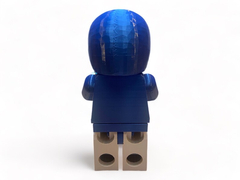 May include: A blue figurine resembling a person, viewed from the back. The figure has a rounded head and a blocky body, with tan-colored feet. The figurine is made of a textured material, and the blue color has a gradient effect.