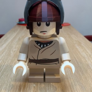 Puede incluir: Una minifigura Lego marr&oacute;n y blanca con un casco azul y marr&oacute;n. La minifigura lleva una t&uacute;nica marr&oacute;n y blanca.