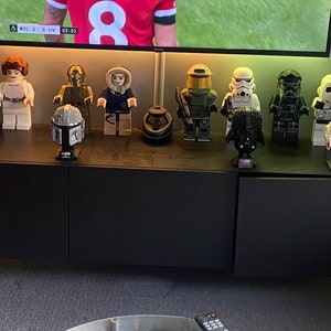 Puede incluir: Una colecci&oacute;n de minifiguras Lego de Star Wars y cascos expuestos en un estante negro. Las minifiguras incluyen a Luke Skywalker, la princesa Leia, C-3PO, Han Solo, un soldado clon, un Stormtrooper, un piloto de caza TIE, un Snowtrooper y Darth Vader. Los cascos incluyen un casco de Boba Fett, un casco de Darth Vader y un casco de Stormtrooper.