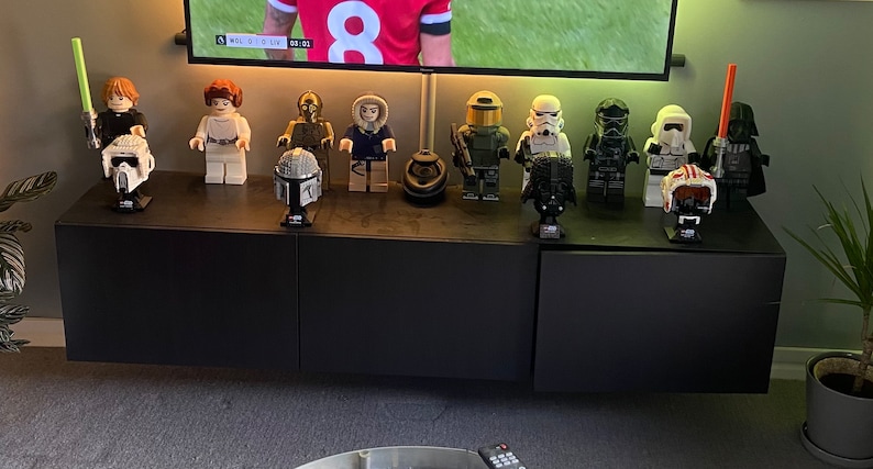 Puede incluir: Una colecci&oacute;n de minifiguras Lego de Star Wars y cascos expuestos en un estante negro. Las minifiguras incluyen a Luke Skywalker, la princesa Leia, C-3PO, Han Solo, un soldado clon, un Stormtrooper, un piloto de caza TIE, un Snowtrooper y Darth Vader. Los cascos incluyen un casco de Boba Fett, un casco de Darth Vader y un casco de Stormtrooper.