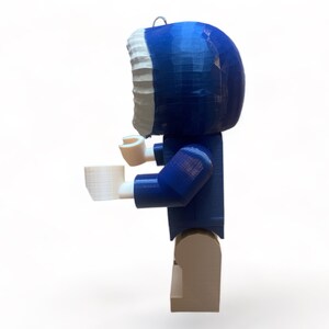 May include: A blue and white figurine of a person wearing a hooded parka, holding a white cup. The figurine is made of plastic and has a block-like design. The figure is standing on a tan base.