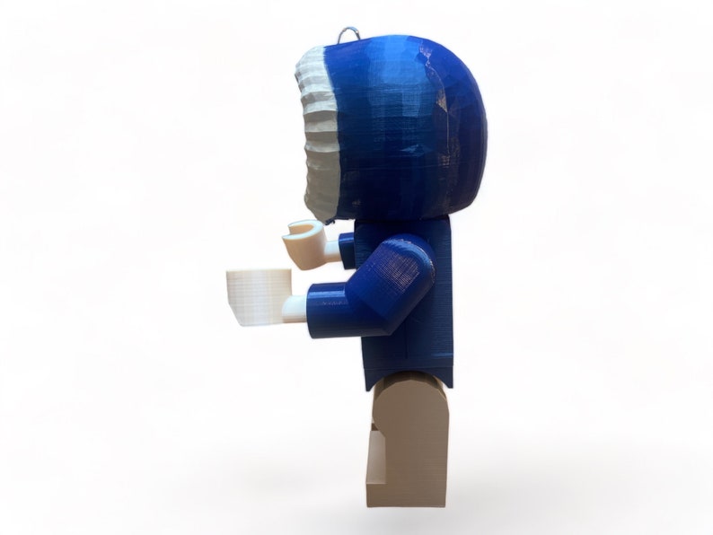 May include: A blue and white figurine of a person wearing a hooded parka, holding a white cup. The figurine is made of plastic and has a block-like design. The figure is standing on a tan base.