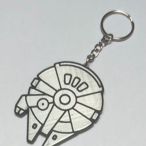 May include: A silver-coloured keychain featuring a flat, stylised representation of a circular spacecraft with a cockpit on the side and a central circular detail. The design includes engraved lines and geometric shapes, attached to a metal ring.