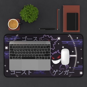 May include: A black desk mat with a purple Gengar design and Japanese text. A laptop, white mouse, and keyboard are on the mat. Other items include a coffee cup, plant, notebook, and pens.