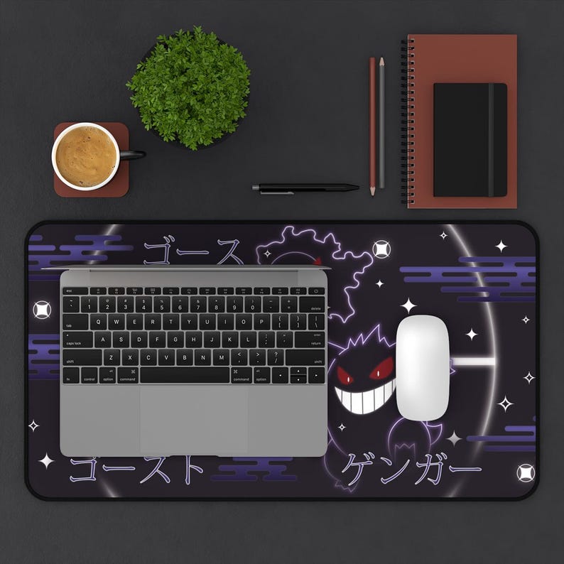 May include: A black desk mat with a purple Gengar design and Japanese text. A laptop, white mouse, and keyboard are on the mat. Other items include a coffee cup, plant, notebook, and pens.