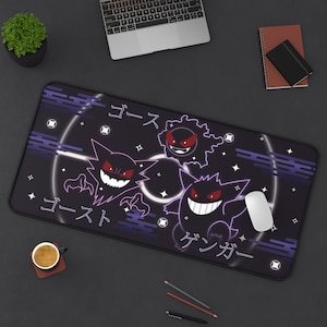 May include: Black rectangular desk mat featuring a purple and white design with three cartoon ghost characters and Japanese text. A white computer mouse is on the mat. The mat is on a black desk.