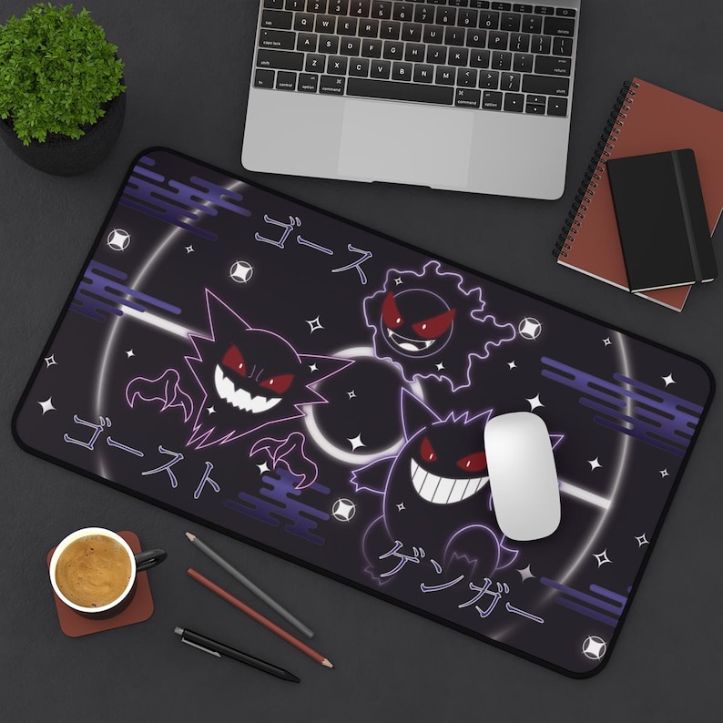 May include: Black rectangular mouse mat featuring a design with Japanese text and cartoon ghost characters in white and purple. A white mouse sits on the mat. A laptop, coffee cup, and notebook are also visible.