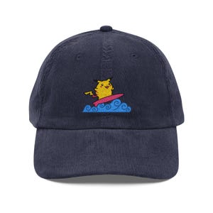 May include: A navy blue corduroy baseball cap featuring an embroidered graphic of a yellow Pikachu character surfing on a pink surfboard atop stylized blue ocean waves. The cap has a structured crown, a curved brim, and a button on the top centre.