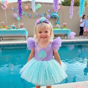 May include: A young girl in a mermaid-themed party dress. The dress has a sequined bodice with a seashell design, tulle sleeves, and a layered, light blue skirt with pearls. She wears a floral headband and lavender shoes. The party has a 'Happy Birthday' banner.