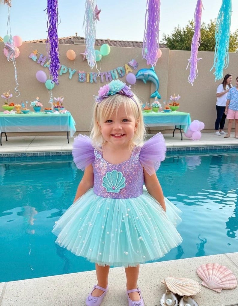May include: A young girl in a mermaid-themed party dress. The dress has a sequined bodice with a seashell design, tulle sleeves, and a layered, light blue skirt with pearls. She wears a floral headband and lavender shoes. The party has a 'Happy Birthday' banner.