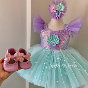 May include: A baby's mermaid-themed outfit set. It includes a lavender and turquoise tutu dress with a sequined bodice and a seashell applique. The set also features matching pink baby shoes with glittery bows and a floral headband. The text "Sparkly Baby Design" is visible.