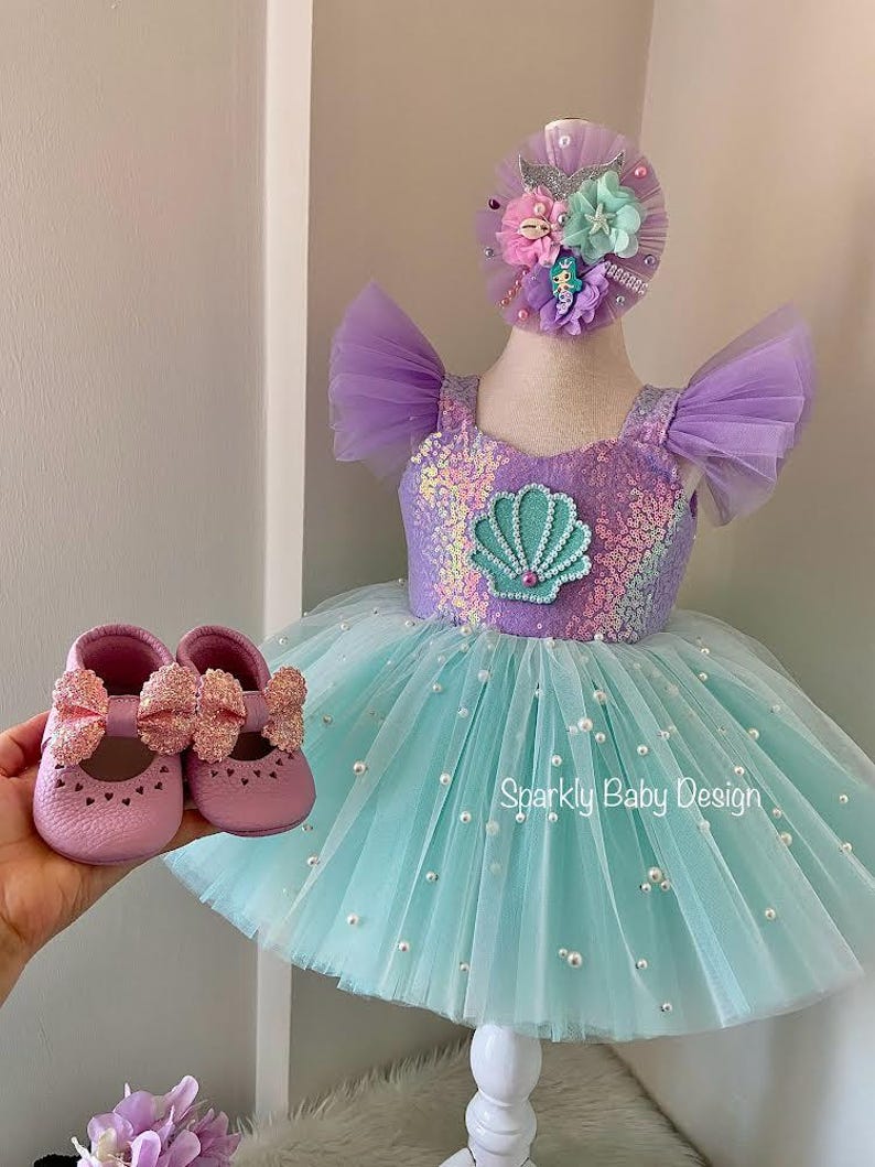 May include: A baby's mermaid-themed outfit set. It includes a lavender and turquoise tutu dress with a sequined bodice and a seashell applique. The set also features matching pink baby shoes with glittery bows and a floral headband. The text "Sparkly Baby Design" is visible.
