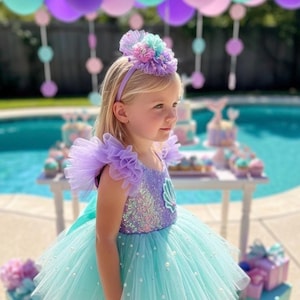 May include: A young girl wearing a mermaid-themed dress with a sequined bodice and a light blue tulle skirt. The dress has ruffled sleeves and a matching headband. The background features balloons and party decorations, creating a festive atmosphere.