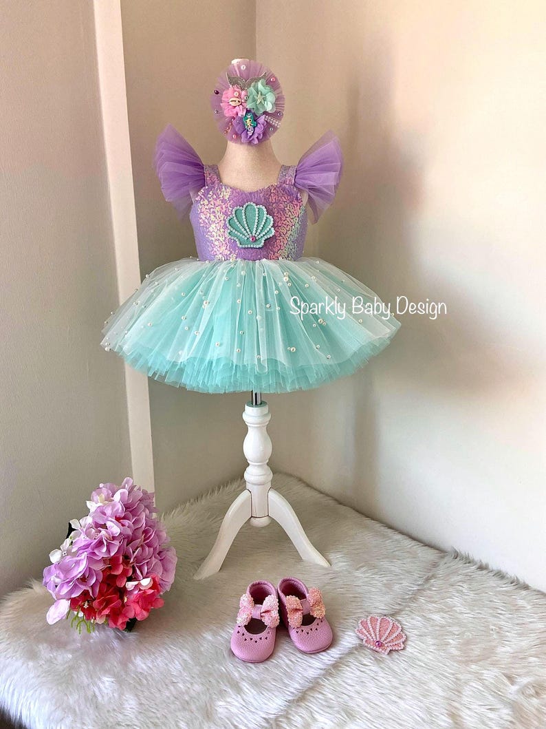 May include: A baby's mermaid-themed outfit, featuring a lavender and turquoise tutu dress with a seashell design, a matching headband, and pink shoes. The dress is displayed on a white mannequin. The text "Sparkly Baby Design" is visible.