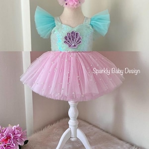 May include: A baby's mermaid-themed dress with a pink tutu skirt and a sequined bodice. The bodice has a seashell design. The dress includes a matching headband and shoes. The text "Sparkly Baby Design" is visible.