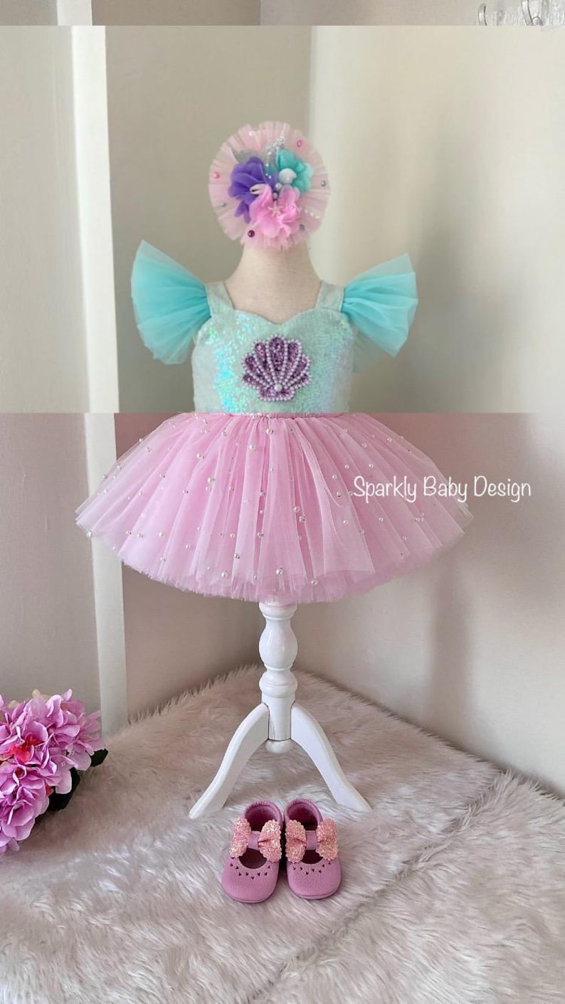 May include: A baby's mermaid-themed dress with a pink tutu skirt and a sequined bodice. The bodice has a seashell design. The dress includes a matching headband and shoes. The text "Sparkly Baby Design" is visible.