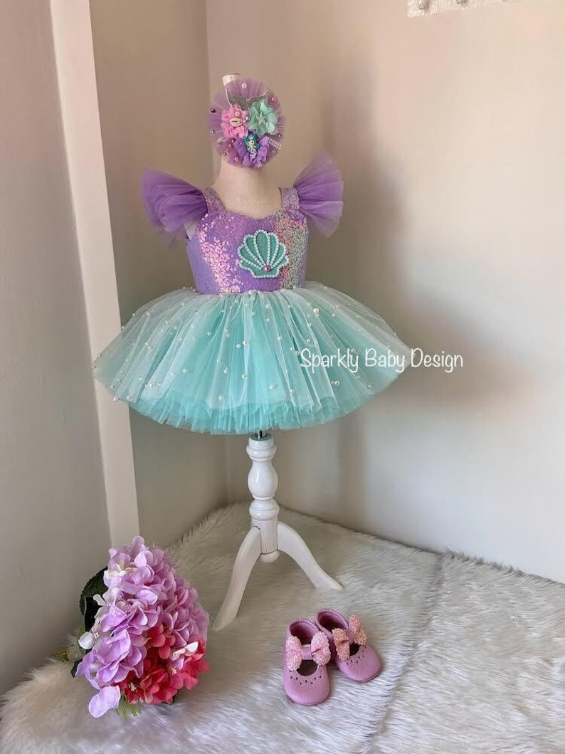 May include: A mermaid-style dress with a purple sequined bodice and a turquoise tulle skirt, decorated with pearls. The dress has a seashell design and is displayed on a white mannequin. A matching headband and pink shoes complete the outfit.
