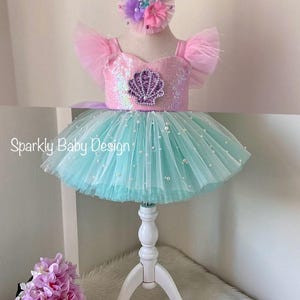 May include: A baby's mermaid-themed outfit, featuring a pink sequined bodice with a seashell design, a turquoise tutu skirt with pearl embellishments, and a floral headband. Pink baby shoes are also included.