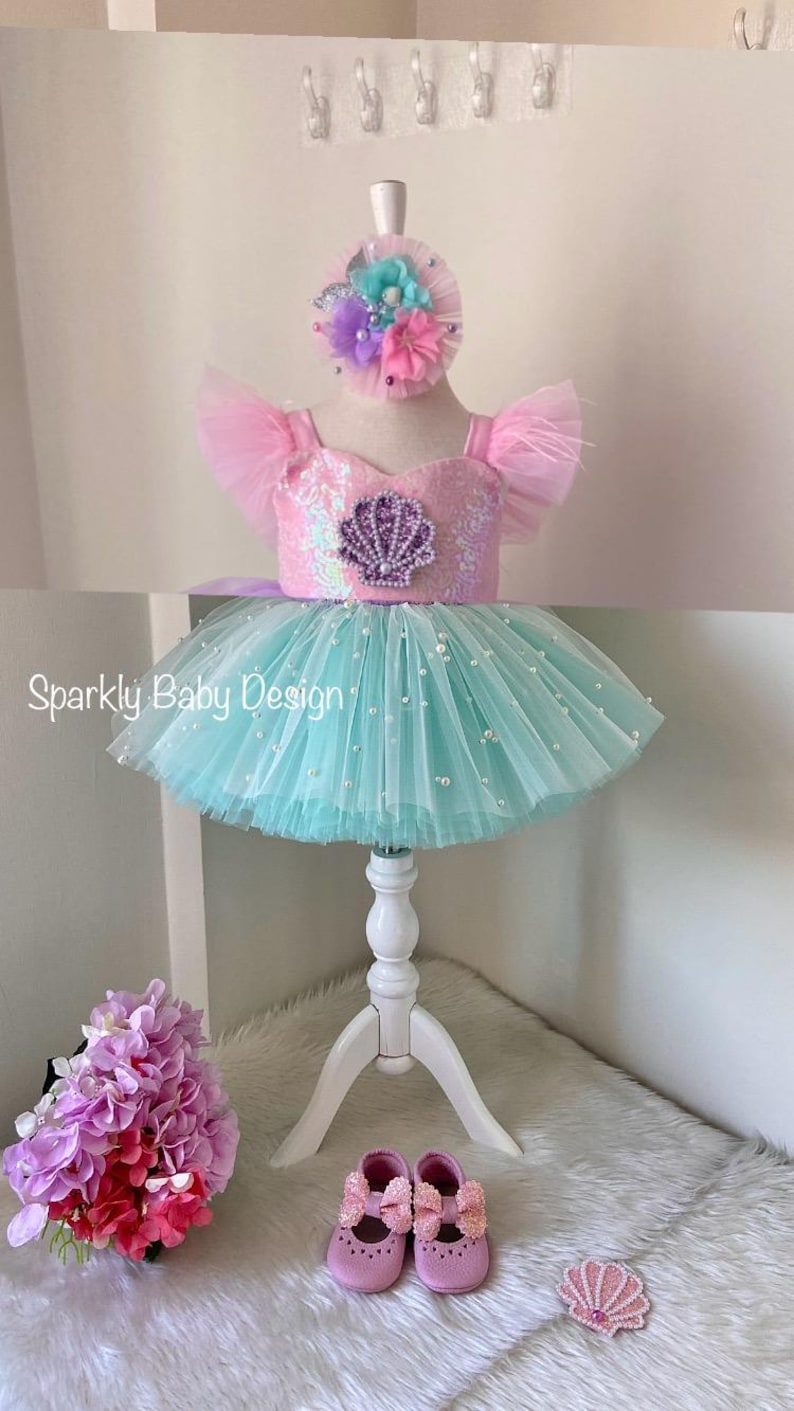 May include: A baby's mermaid-themed outfit, featuring a pink sequined bodice with a seashell design, a turquoise tutu skirt with pearl embellishments, and a floral headband. Pink baby shoes are also included.