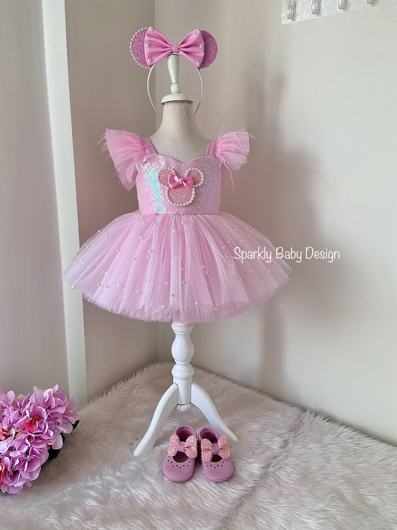 May include: Pink tulle dress with a sequined bodice featuring a pink and white Mickey Mouse head design. The dress is adorned with pearl-like embellishments and comes with a matching pink headband with Mickey Mouse ears. The dress is displayed on a white mannequin and is accompanied by a pair of pink shoes with bows.