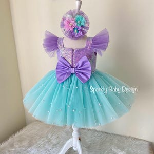 May include: A baby girl's dress and matching headband. The dress has a purple sequined bodice, a large purple bow, and a turquoise tulle skirt with pearls. The headband is decorated with flowers.