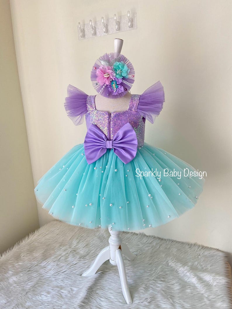 May include: A baby girl's dress and matching headband. The dress has a purple sequined bodice, a large purple bow, and a turquoise tulle skirt with pearls. The headband is decorated with flowers.
