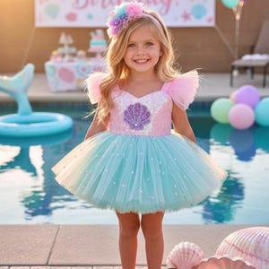 May include: A young girl in a mermaid-themed birthday outfit. The dress is pink and turquoise with a seashell design. She wears a matching headband and shoes. A banner that says "Happy Birthday!" is in the background.