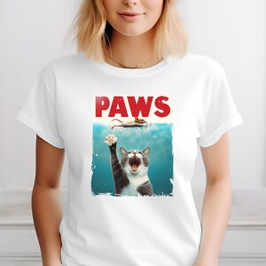 May include: White t-shirt with a graphic of a cat reaching out of the water with the text "PAWS" above it.