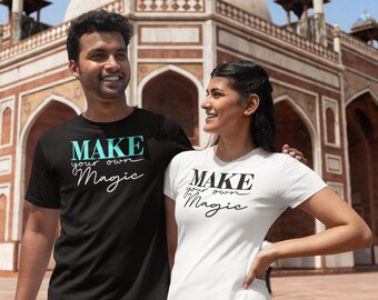 May include: Two people wearing matching t-shirts with the text "MAKE your own Magic" in black and turquoise lettering. The person on the left is wearing a black t-shirt and the person on the right is wearing a white t-shirt. They are standing in front of a red brick building with arches.