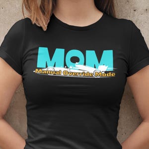 Puede incluir: Camiseta negra con la palabra "MOM" en letras turquesas grandes. Debajo, el texto "-Manual Override Mode" y un dibujo de un coche de carreras blanco.