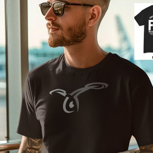 May include: A black t-shirt with a gray stylized logo of a drone on the front. The back of the shirt has the text "CERTIFIED REMOTE PILOT FAA" in white. The shirt is being worn by a person wearing sunglasses.