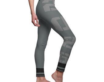 May include: Gray leggings with a large, lighter gray geometric design on the legs. The leggings have a black band at the waist and at the ankles. The leggings are form-fitting and appear to be made of a stretchy material.