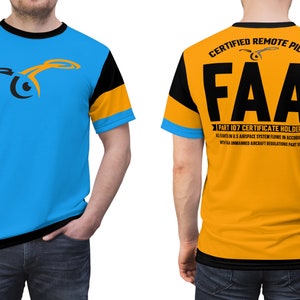 Peut inclure: Un t-shirt bleu, noir et orange avec un graphique de drone sur le devant et le texte "CERTIFIED REMOTE PILOT FAA PART 107 CERTIFICATE HOLDER" au dos.