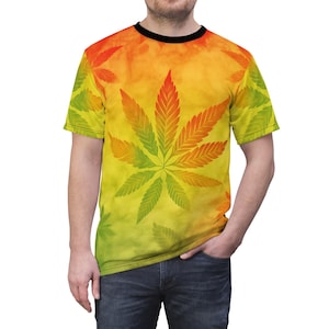 Könnte beinhalten: Ein kurzärmeliges T-Shirt mit einem großen Cannabisblatt-Design. Das Blatt wechselt von Grün zu Orange, vor einem gelb-orangen Batik-Hintergrund. Das Shirt hat einen schwarzen Kragen und wird von einer Person getragen.