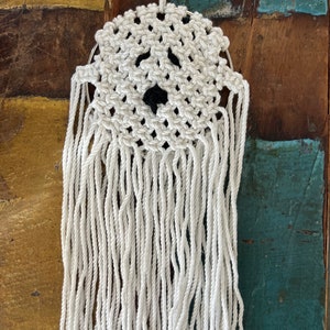 Könnte beinhalten: Weißer Makramee-Geister-Wandbehang mit schwarzen Augen. Der Geist ist aus weißem Garn gefertigt und hängt an einer Holzoberfläche.