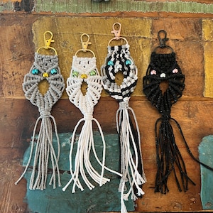 Puede incluir: Cuatro llaveros de búho de macramé con borlas de diferentes colores. Los búhos están hechos con hilo gris, blanco, negro y blanco a rayas y negro. Cada búho tiene una pequeña campana en el pecho.