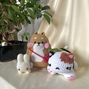 Op de afbeelding: Drie gehaakte knuffels: een bruine en witte hond, een wit konijn en een witte kat met bruine en oranje accenten. De kat draagt een groene en gele sushirol.