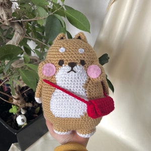 Op de afbeelding: Een gehaakt bruin en wit honden knuffeldier met roze wangen en een rode schoudertas.