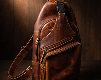 Puede incluir: Una mochila bandolera de cuero marrón con múltiples compartimentos con cremallera, herrajes de metal en tono latón antiguo y una hebilla de plástico negra en la correa ajustable. La superficie muestra una textura natural y desgastada.