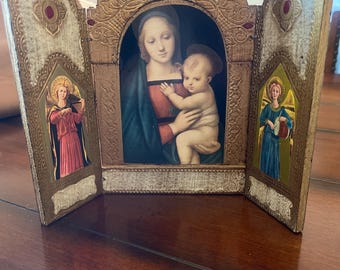 Pode incluir: Um tríptico de madeira com uma moldura dourada e três painéis. O painel central apresenta uma pintura da Virgem Maria segurando o menino Jesus. Os painéis laterais apresentam pinturas de anjos.