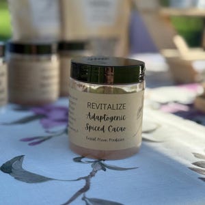 May include: A clear jar with a black lid, labeled "Revitalize Adaptogenic Spiced Cacao" from Forest Moon Medicine, sits on a floral tablecloth. Other jars and bags are visible in the background, suggesting a health food product.