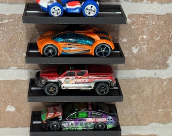 Op de afbeelding: Vijf speelgoedauto's tentoongesteld op een zwart wandrek met een zwart spoor. De auto's zijn een rode Mario Kart, een oranje sportwagen, een rode pick-up truck, een zwarte en paarse auto met de tekst "Save Our Souls" erop en een rode en gouden sportwagen.
