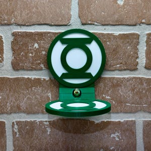 May include: Green Lantern logo wall mount with a shelf. The logo is white with a green outline and is mounted on a green base. The shelf is also green and has a slight curve.