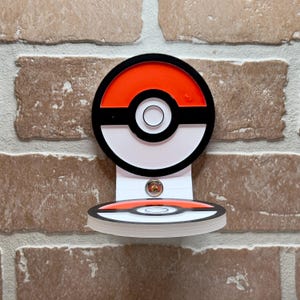 May include: A black, white, and red Pokeball wall mount with a round base. The Pokeball is a popular symbol from the Pokemon franchise.