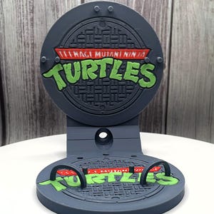 May include: A gray 3D-printed stand featuring the Teenage Mutant Ninja Turtles logo. The design includes a manhole cover with the words "Teenage Mutant Ninja Turtles" in green and red. The stand has a circular base and a vertical support.