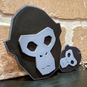 May include: Black and gray gorilla face-shaped wall decor and keychain. The gorilla face has a gray face and a black outline. The keychain has a silver keyring. The items are on a dark surface.