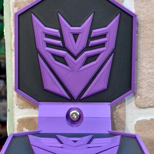 May include: Purple and black wall mount with a 3D printed Decepticon symbol. The mount has a shelf that folds down.