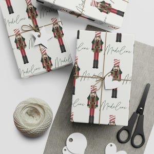 Puede incluir: Papel de regalo blanco con un patrón repetido de un soldado cascanueces en rojo, verde y negro. La palabra "Madeline" está escrita en cursiva en el papel. Los regalos envueltos están atados con hilo y tienen etiquetas en forma de estrella y corazón.