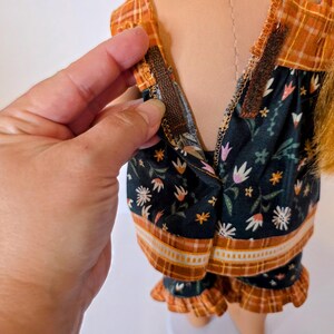 May include: A doll wearing a two-piece pajama set. The top and shorts are dark green with a floral pattern and orange trim. The top has a plaid orange yoke. A hand is shown opening the zipper on the top.
